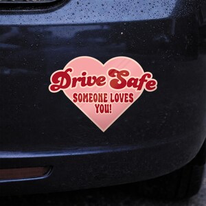 May include: A pink heart-shaped bumper sticker with the words "Drive Safe" in red script and "SOMEONE LOVES YOU!" in a smaller font. The sticker is affixed to a dark-colored vehicle.