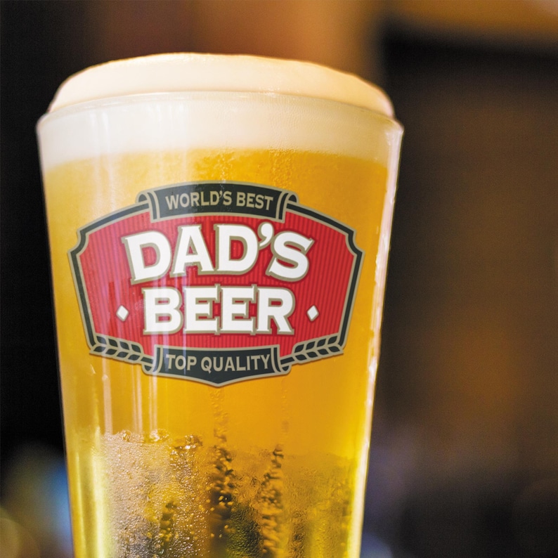 May include: A glass of beer with a foamy head. The glass has a label that reads "WORLD'S BEST DAD'S BEER TOP QUALITY" in red and white. The beer is a golden color.