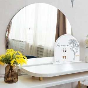 May include: White jewelry stand with the name Monica written on it. The stand has a floral design and is used to display earrings. The stand is on a white vanity table with a round mirror behind it.