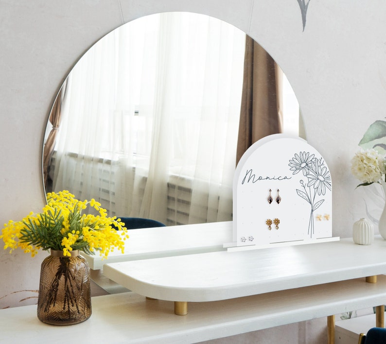 May include: White jewelry stand with the name Monica written on it. The stand has a floral design and is used to display earrings. The stand is on a white vanity table with a round mirror behind it.