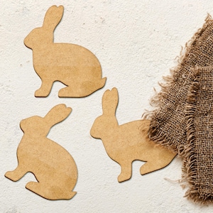 May include: Three wooden rabbit silhouettes on a white background. The rabbits are cut out of brown wood and are arranged in a triangular pattern. A piece of burlap fabric is in the upper right corner of the image.