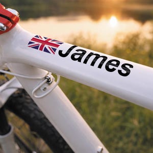 May include: White bicycle frame with a Union Jack sticker and the name "James" in black lettering. The bike has a red component and a black tire. The background shows a blurred view of a body of water and a sunset.