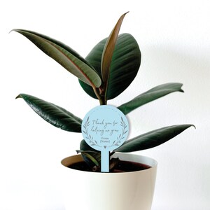 May include: A light blue plant marker with the text "Thank you for helping me grow. From [Name]" in gray lettering. The marker is in the shape of a circle with a wreath design around the text. The marker is inserted into a potted plant.