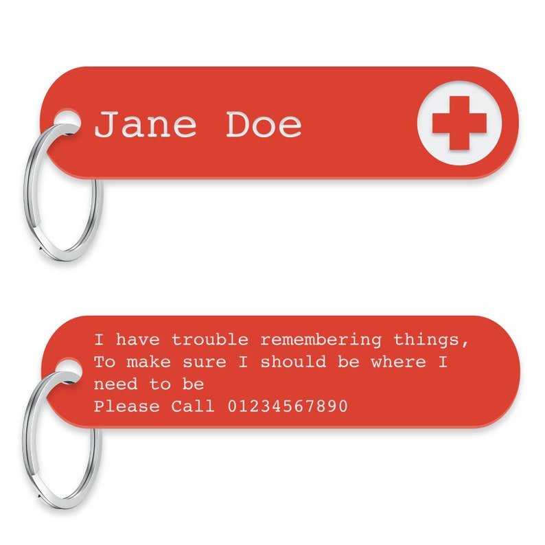 K&ouml;nnte beinhalten: Roter Schl&uuml;sselanh&auml;nger mit einem wei&szlig;en Kreuz und dem Text "Jane Doe" oben. Der untere Teil des Schl&uuml;sselanh&auml;ngers tr&auml;gt den Text "I have trouble remembering things, To make sure I should be where I need to be Please Call 01234567890".