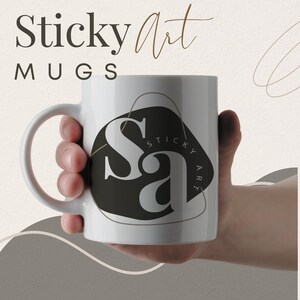 May include: White ceramic mug with a black and gold logo that says "Sticky Art" in a stylized font. The mug is being held by a hand.