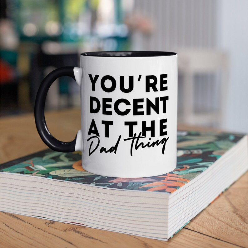 May include: White ceramic mug with a black handle and black rim. The mug has black text that reads "You're Decent at the Dad Thing."