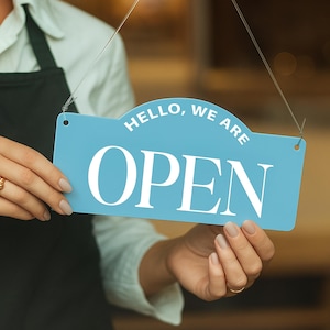 May include: A light blue sign with the words "HELLO, WE ARE OPEN" in white lettering. The sign is held by a person. The sign is rectangular with a curved top and is suspended by thin wires.