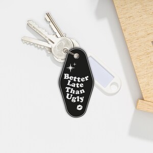 May include: Black motel key chain with the text "Better Late Than Ugly" in white with a small star and a lip print. The keychain is attached to a silver key.