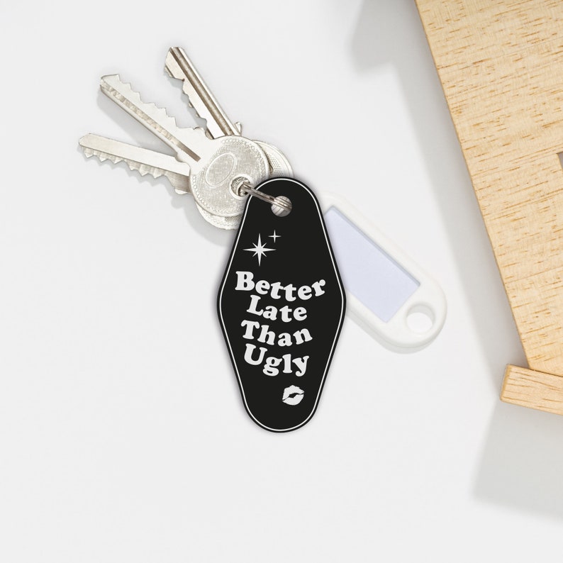 May include: Black motel key chain with the text "Better Late Than Ugly" in white with a small star and a lip print. The keychain is attached to a silver key.