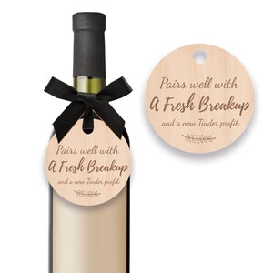 May include: A wine bottle with a black ribbon and a wooden tag that reads "Pairs well with A Fresh Breakup and a new Tinder profile." A second wooden tag with the same text is also visible.