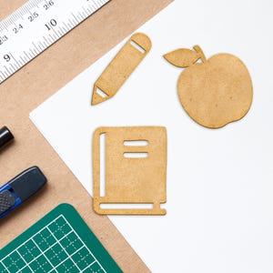 K&ouml;nnte beinhalten: Ein wei&szlig;es Blatt Papier mit einem braunen Bleistift, einem braunen Buch und einem braunen Apfel, die aus Papier ausgeschnitten sind. Das Papier liegt auf einer braunen Oberfl&auml;che.