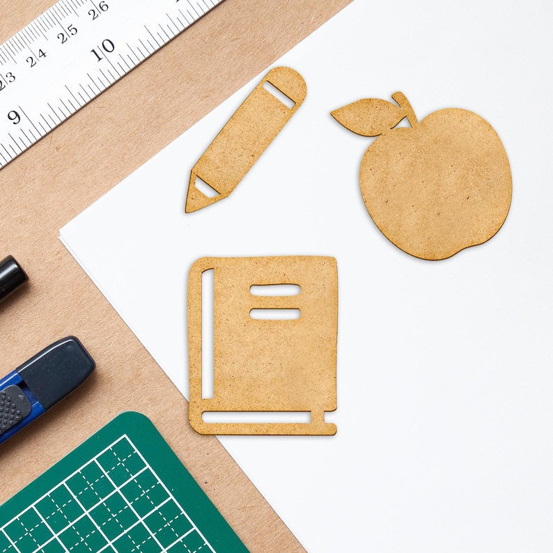 K&ouml;nnte beinhalten: Ein wei&szlig;es Blatt Papier mit einem braunen Bleistift, einem braunen Buch und einem braunen Apfel, die aus Papier ausgeschnitten sind. Das Papier liegt auf einer braunen Oberfl&auml;che.