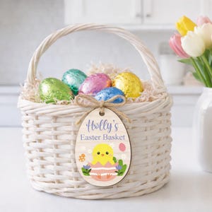 May include: A white wicker Easter basket filled with colorful foil-wrapped eggs and decorative paper. A wooden egg-shaped tag with the text "Holly's Easter Basket" and a chick illustration hangs from the basket. A bouquet of tulips is in the background.