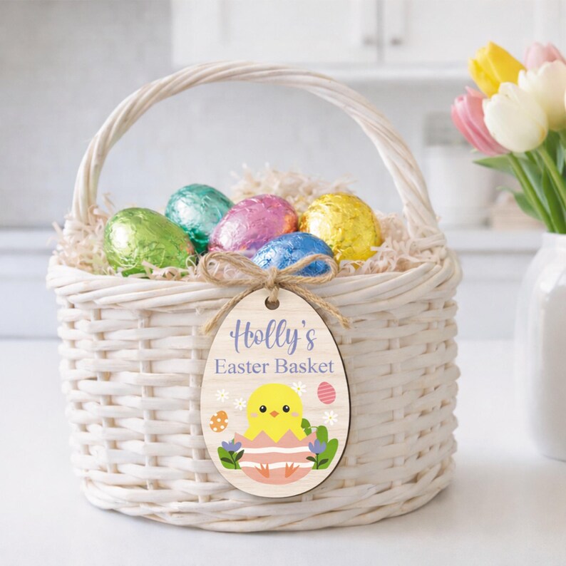 May include: A white wicker Easter basket filled with colorful foil-wrapped eggs and decorative paper. A wooden egg-shaped tag with the text "Holly's Easter Basket" and a chick illustration hangs from the basket. A bouquet of tulips is in the background.