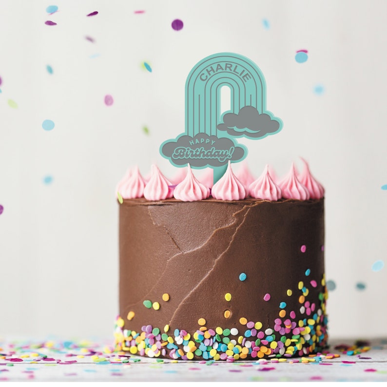 Op de afbeelding: Een chocoladetaart met roze glazuur en kleurrijke sprinkles. Een turquoise regenboogtaarttopper met de naam "Charlie" en de woorden "Happy Birthday!" staat bovenop de taart.