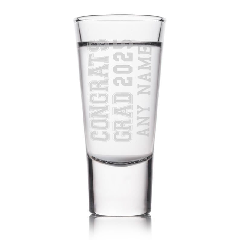 May include: A clear shot glass with the words "CONGRATS GRAD 2023 ANY NAME" etched in a bold, sans-serif font. The glass is filled with a clear liquid, and the design is centered on the glass.