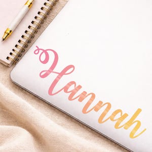 May include: A white laptop with the name "Hannah" in a gradient of pink, orange, and yellow script. A gold-tipped pen and a spiral notebook are in the upper left corner. The laptop rests on a beige fabric.