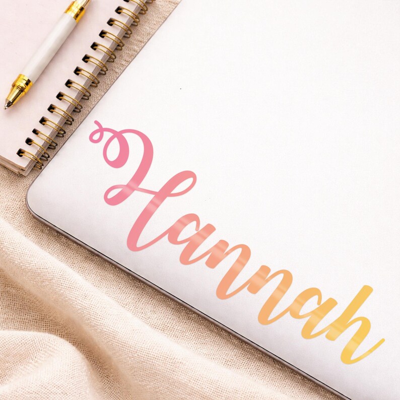 May include: A white laptop with the name "Hannah" in a gradient of pink, orange, and yellow script. A gold-tipped pen and a spiral notebook are in the upper left corner. The laptop rests on a beige fabric.