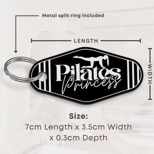 以下が含まれることがあります： 金属製のスプリットリングが付いた、白と黒のピラティスプリンセスのキーホルダー。キーホルダーのサイズは長さ7cm、幅3.5cm、厚さ0.3cmです。「Pilates Princess」の文字とピラティスのポーズがデザインされています。