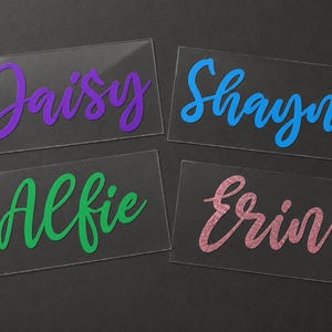 May include: Four clear acrylic name tags with names in various colors. "Daisy" is in purple, "Shayne" in blue, "Alfie" in green, and "Erin" in pink glitter. The names are in a cursive font.