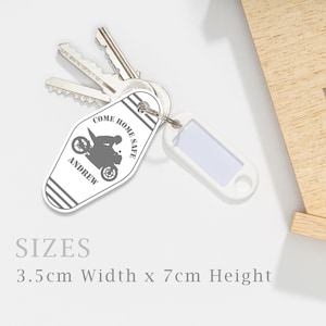 Op de afbeelding: Witte sleutelhanger met een motorfietsafbeelding en de tekst "COME HOME SAFE ANDREW". De sleutelhanger is bevestigd aan sleutels en een wit label. Afmetingen: 3,5 cm breed x 7 cm hoog.