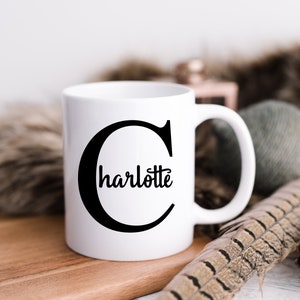 K&ouml;nnte beinhalten: Eine wei&szlig;e Keramiktasse mit einem schwarzen Buchstaben "C" und dem Namen "Charlotte" innerhalb des Buchstabens. Die Tasse steht auf einer Holzoberfl&auml;che.
