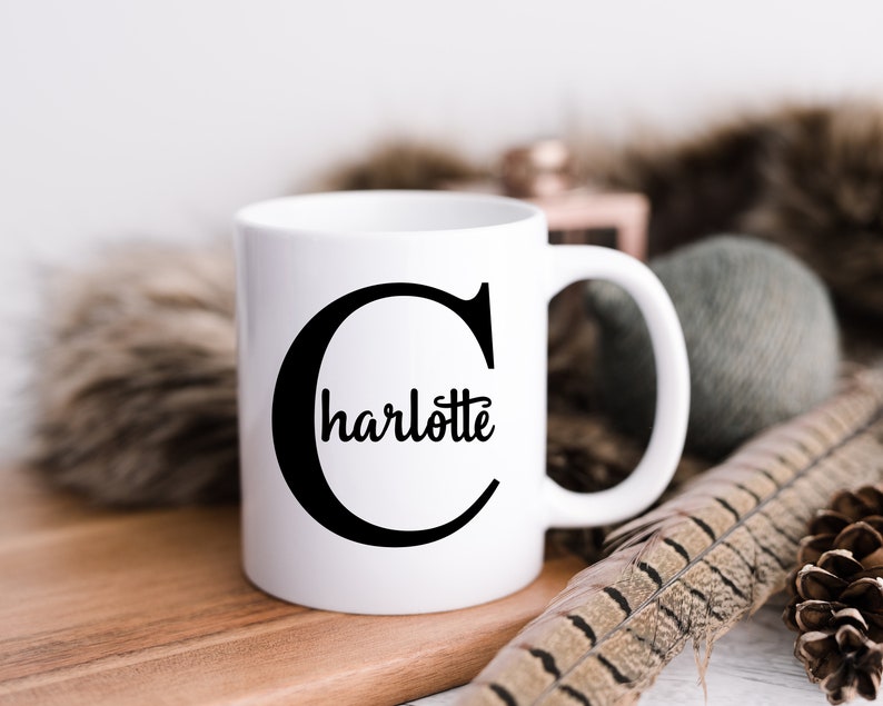K&ouml;nnte beinhalten: Eine wei&szlig;e Keramiktasse mit einem schwarzen Buchstaben "C" und dem Namen "Charlotte" innerhalb des Buchstabens. Die Tasse steht auf einer Holzoberfl&auml;che.