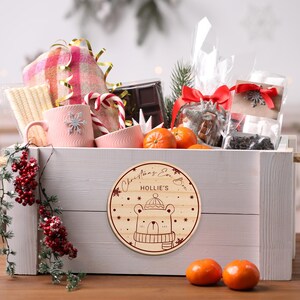 May include: A white wooden box filled with Christmas gifts, including a pink mug, a blanket, candy canes, chocolate, and oranges. The box has a round wooden tag attached to it that says "Christmas Eve Box Hollie's" and features a cartoon bear wearing a hat and scarf.