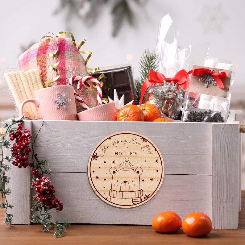 May include: A white wooden box filled with Christmas gifts, including a pink mug, a blanket, candy canes, chocolate, and oranges. The box has a round wooden tag attached to it that says "Christmas Eve Box Hollie's" and features a cartoon bear wearing a hat and scarf.