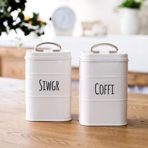 May include: Two white metal canisters with black lettering. The canister on the left says "Sugar" and the canister on the right says "Coffee".