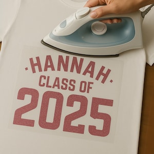 May include: A white t-shirt with a heat transfer design that reads "HANNAH CLASS OF 2025" in a red glitter font. An iron is being used to apply the design to the shirt.