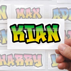 Op de afbeelding: Een sticker met de naam "KIAN" in een graffiti-stijl. De letters zijn gearceerd met een verloop van groen naar geel, omlijnd in zwart. De sticker wordt in een hand gehouden, met andere naamstickers op de achtergrond.
