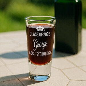 May include: A clear shot glass filled with a dark liquid, featuring the text "CLASS OF 2025 George BSC PSYCHOLOGY" in white. A graduation cap graphic is above the name. A dark green bottle is in the background.