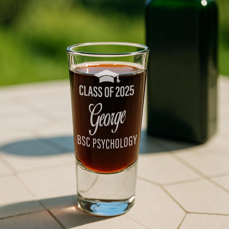 May include: A clear shot glass filled with a dark liquid, featuring the text "CLASS OF 2025 George BSC PSYCHOLOGY" in white. A graduation cap graphic is above the name. A dark green bottle is in the background.
