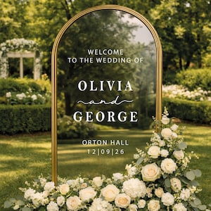 Op de afbeelding: Een gouden trouwbord in de vorm van een boog met de tekst "WELCOME TO THE WEDDING OF OLIVIA and GEORGE" en de datum "12|09|26". Versierd met witte rozen en groen, tegen een groene grasmat.