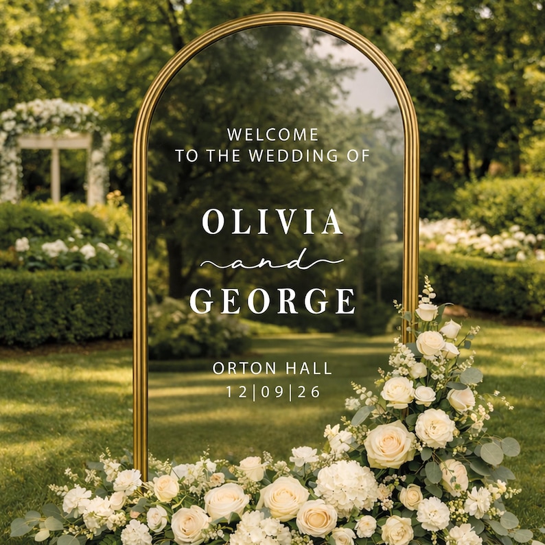 Op de afbeelding: Een gouden trouwbord in de vorm van een boog met de tekst "WELCOME TO THE WEDDING OF OLIVIA and GEORGE" en de datum "12|09|26". Versierd met witte rozen en groen, tegen een groene grasmat.