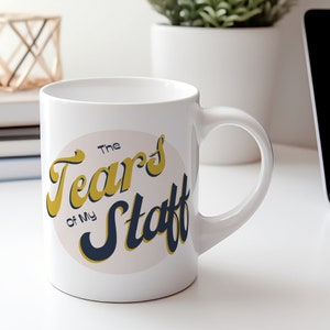 May include: White ceramic mug with a gold and blue design that says "The Tears of My Staff" in a retro font.