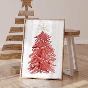 May include: A framed watercolor print of a red Christmas tree with a star on top and string lights. The artwork is in a wooden frame and leans against a wall. A wooden Christmas tree and a small stool are in the background.