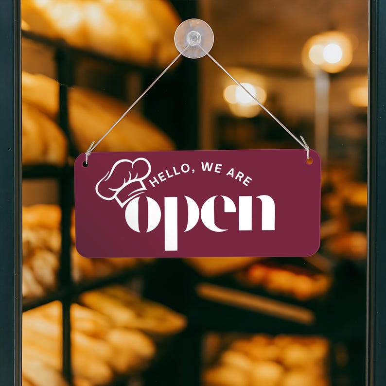Peut inclure: Une enseigne rectangulaire bordeaux avec les mots "Hello, we are open" en blanc, ainsi qu'un dessin de toque de chef. L'enseigne est suspendue &agrave; une porte vitr&eacute;e avec des produits de boulangerie en arri&egrave;re-plan.