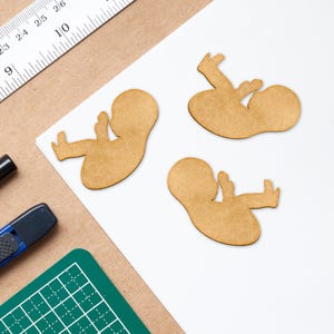 May include: Three wooden cutouts of a fetus in a fetal position. The cutouts are light brown and are on a white surface. A ruler, a utility knife, and a cutting mat are in the background.