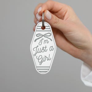K&ouml;nnte beinhalten: Ein wei&szlig;-grauer Schl&uuml;sselanh&auml;nger mit dem Text "I'm Just a Girl" in einer Schreibschrift. Der Schl&uuml;sselanh&auml;nger hat ein Retro-Motel-Schl&uuml;ssel-Design mit einer Schleife und gestreiften Details. Ein silberner Schl&uuml;sselring ist befestigt.