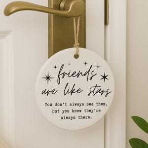 May include: A round, white ceramic hanging ornament with the text "friends are like stars." The ornament is suspended by a twine string and hangs from a gold-colored door handle. The text includes the quote: "You don't always see them, but you know they're always there."