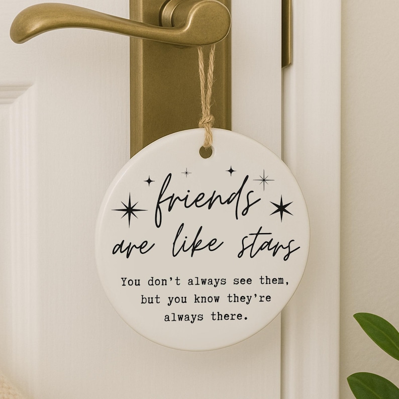 May include: A round, white ceramic hanging ornament with the text "friends are like stars." The ornament is suspended by a twine string and hangs from a gold-colored door handle. The text includes the quote: "You don't always see them, but you know they're always there."