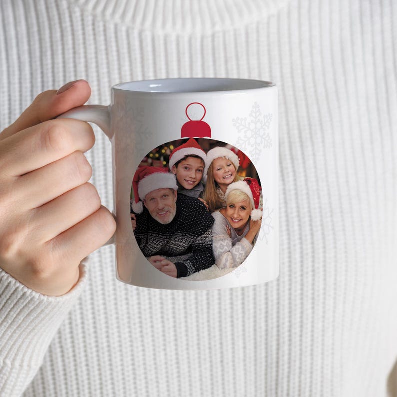 May include: White ceramic mug featuring a family photo inside a Christmas ornament design. The ornament is red and white, with a family of four wearing Santa hats. Snowflake accents are on the mug.