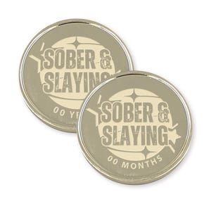 Puede incluir: Dos monedas redondas de color plateado con fondo blanco. Las monedas tienen el texto "SOBER & SLAYING" en negro con un aspecto desgastado. Una moneda dice "00 A&Ntilde;OS" y la otra "00 MESES".