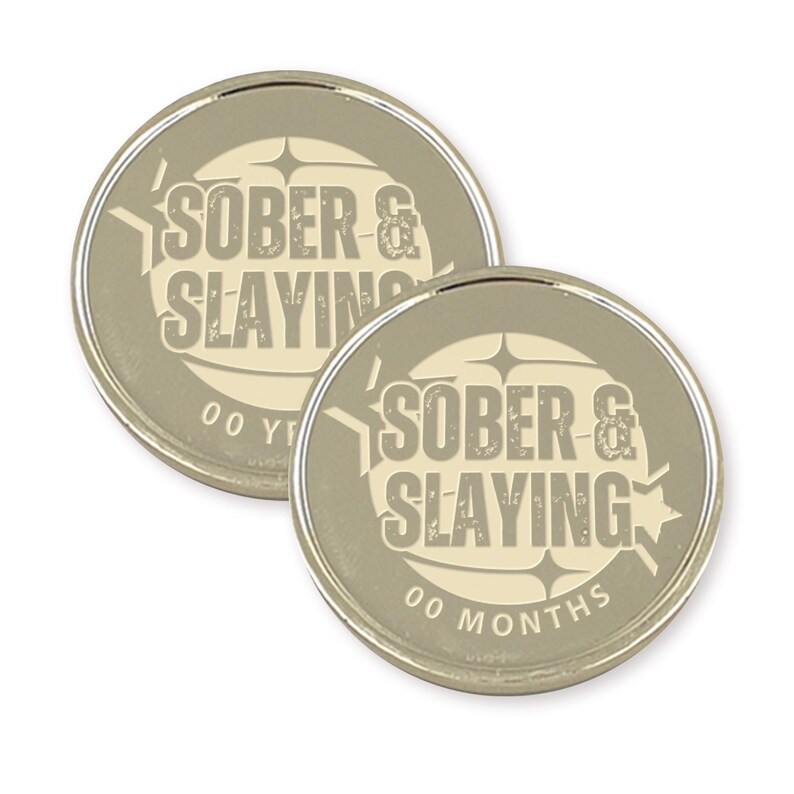Puede incluir: Dos monedas redondas de color plateado con fondo blanco. Las monedas tienen el texto "SOBER & SLAYING" en negro con un aspecto desgastado. Una moneda dice "00 A&Ntilde;OS" y la otra "00 MESES".