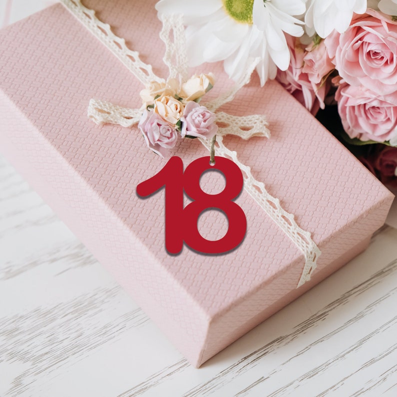 May include: A pink gift box adorned with a white lace ribbon and a bouquet of pink and white flowers. A red number 18 tag hangs in front of the box, suggesting a birthday or special occasion. The box sits on a white wooden surface.