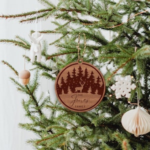 Op de afbeelding: Een ronde houten kerstversiering met een lasergesneden ontwerp van een bos sc&egrave;ne met een hert, maan en sterren. De versiering is gepersonaliseerd met de tekst "The Jones Family".