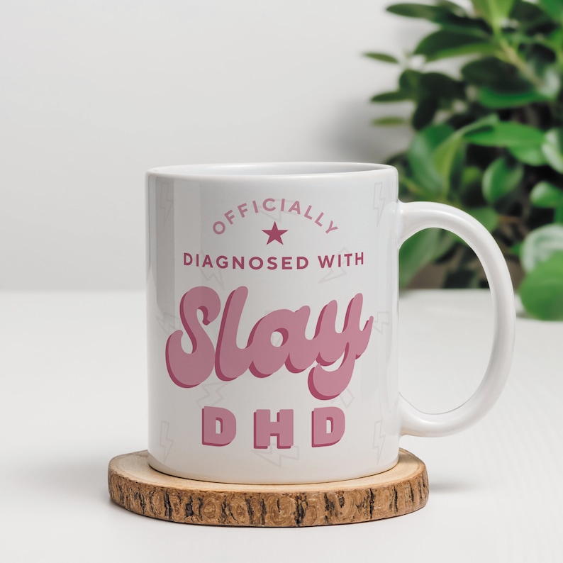 May include: White ceramic mug with a pink design that says "Officially Diagnosed with Slay DHD". The mug has a star and a pattern of small lightning bolts.