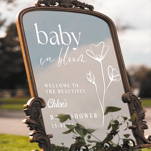 May include: A gold-framed mirror with a white inscription that reads "baby in bloom" and "Welcome to the beautiful Chloe's Baby Shower 15/01/24". The mirror is decorated with a white floral design.